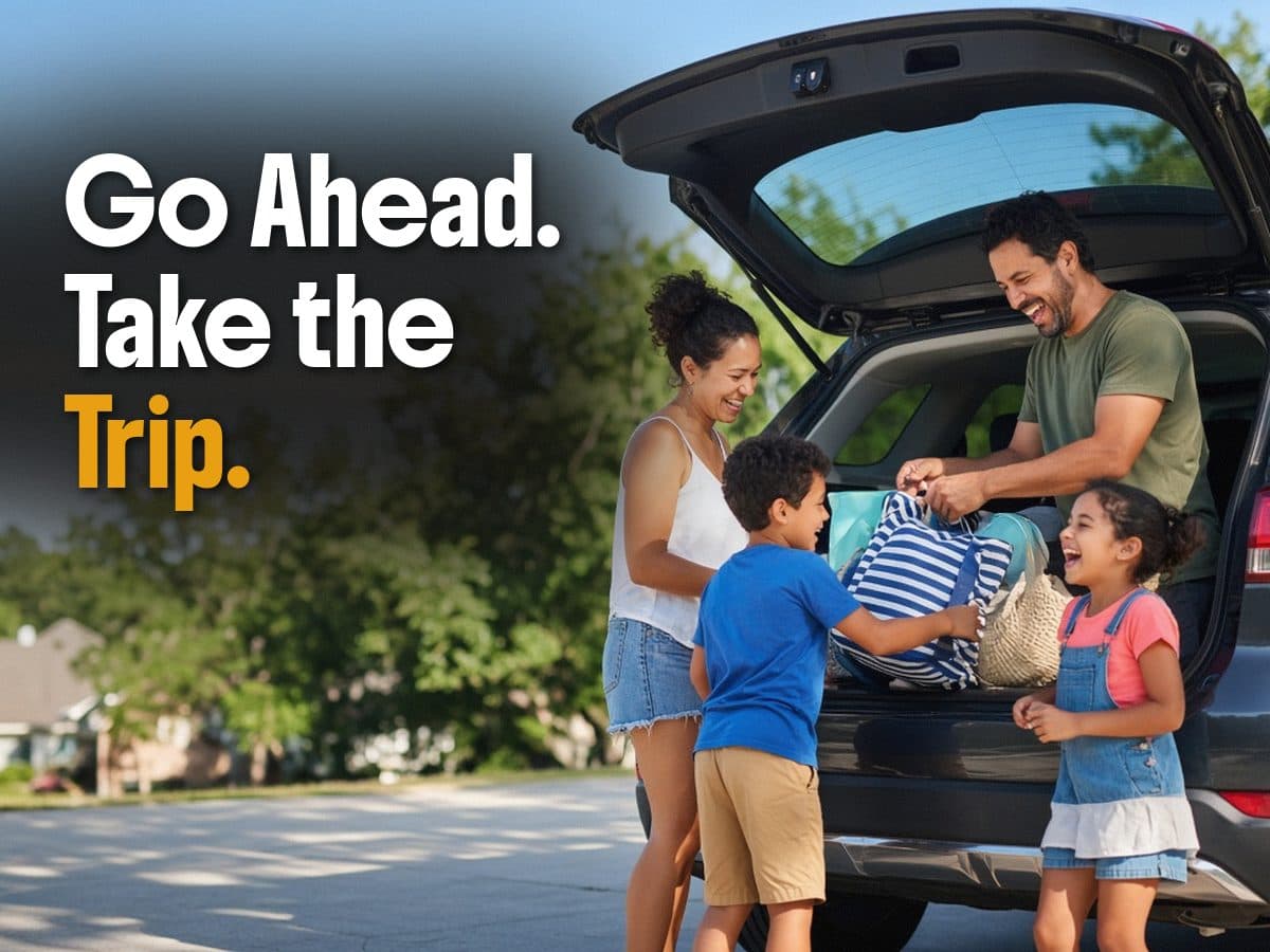 A family of four packs bags into an open SUV on a sunny street, under the words Go Ahead. Take the Trip.