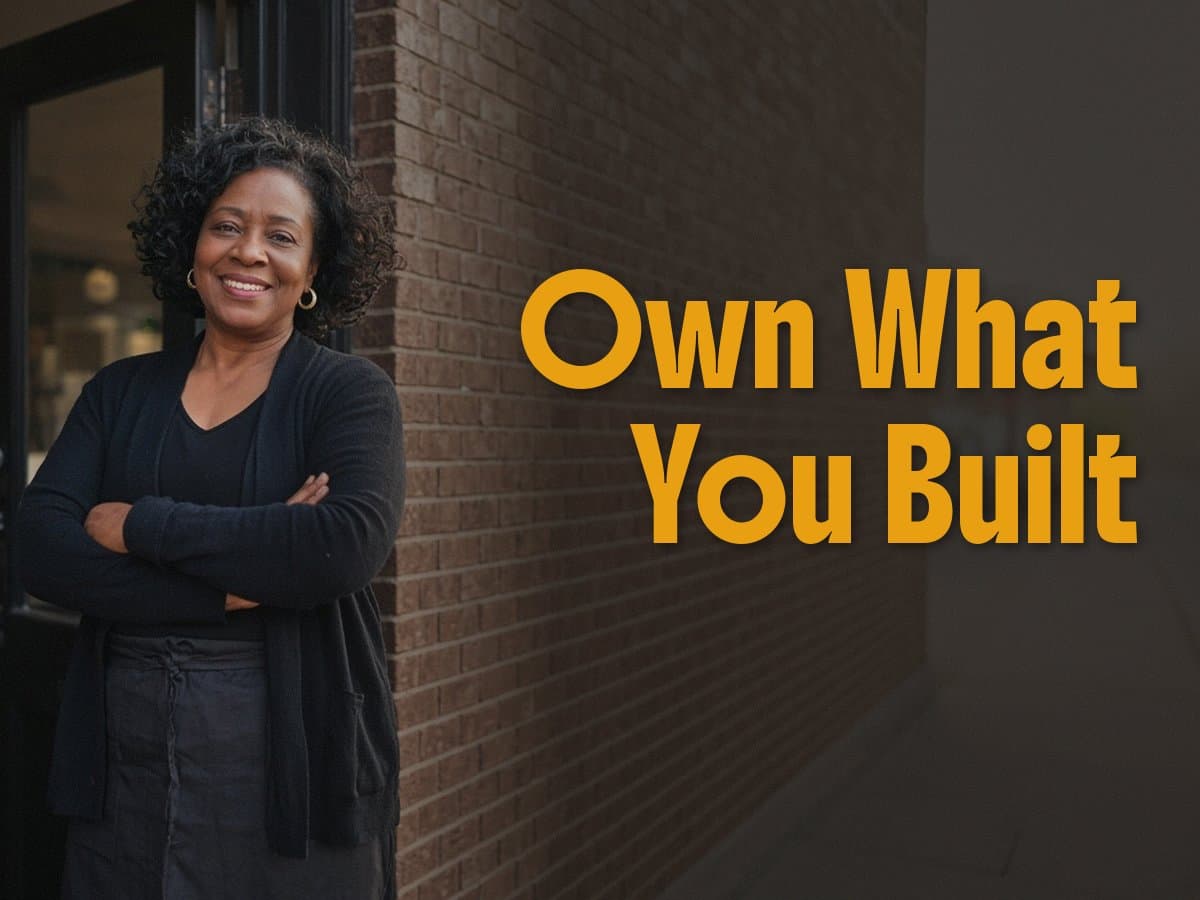 A smiling woman with crossed arms stands by a brick doorway, next to the gold words Own What You Built.